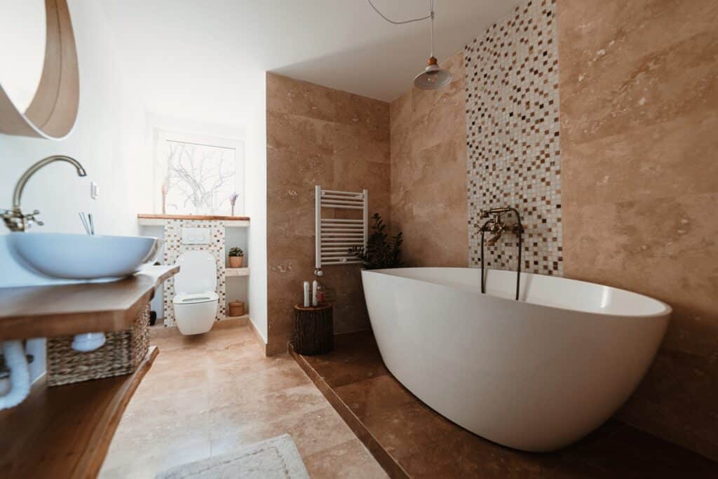 Contemporary bathroom with a white freestanding bathtub, beige tiled walls, and a wall-mounted toilet near a mosaic tile accent wall.